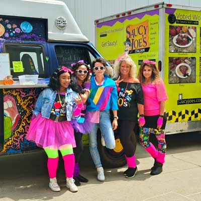 Women smiling in front of Saucy Joe's and Good Vibrations truck at company party